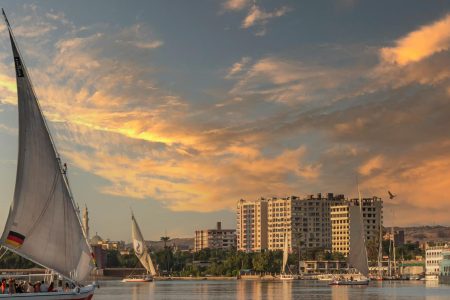 A traditional felucca sails the Nile at sunset with cityscape in the background.