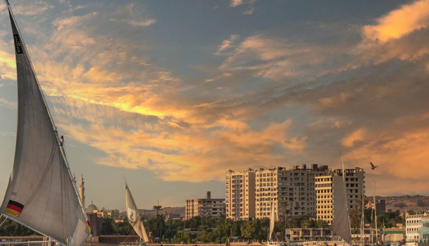 A traditional felucca sails the Nile at sunset with cityscape in the background.
