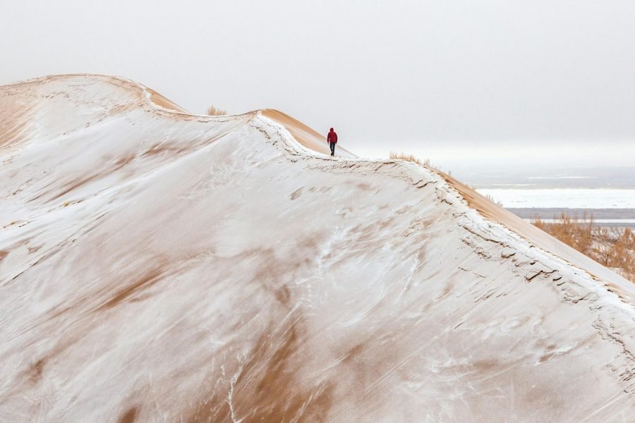 A lone hiker walks along snow-covered sand dunes in a vast, snowy landscape.
